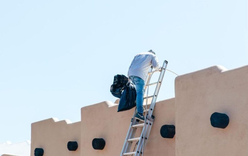 Man climbs onto an Arizona rooftop for repair work, with safety gear and tools.